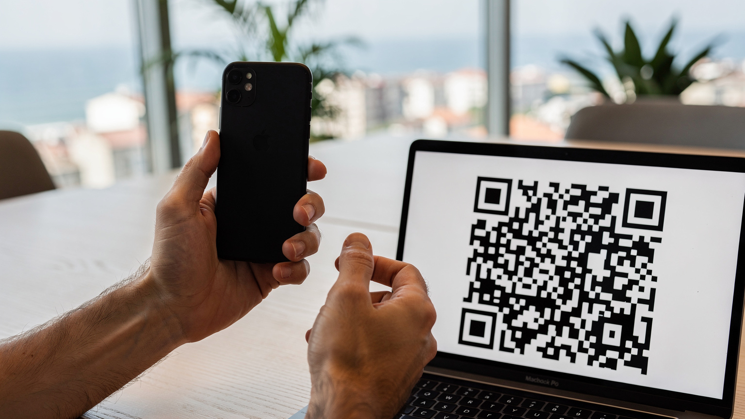 Близък план на ръце, които държат смартфон и сканират QR код от друг екран, за да инсталират eSIM, с разфокусиран фон на модерен интериор.