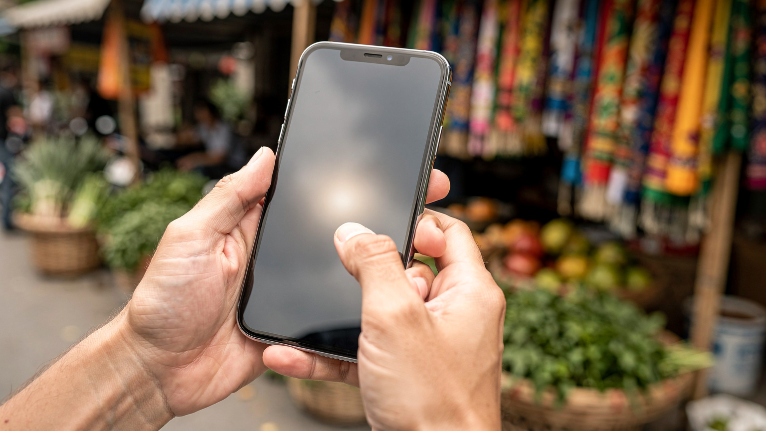 A close-up of a traveller's hands tapping on their smartphone screen, checking data usage within an eSIM app, with a colourful market stall blurred in the background, suggesting they are in a vibrant street market.
