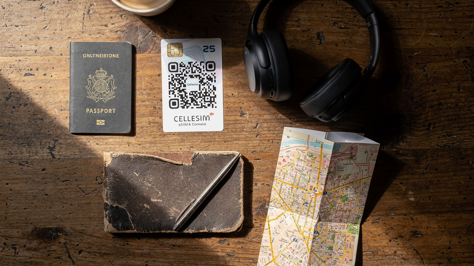An overhead flat-lay shot of travel essentials, including a passport, a Cellesim eSIM QR card, noise-canceling headphones, a small travel journal, and a carefully folded map, all set on a rustic wooden table in a cafe.