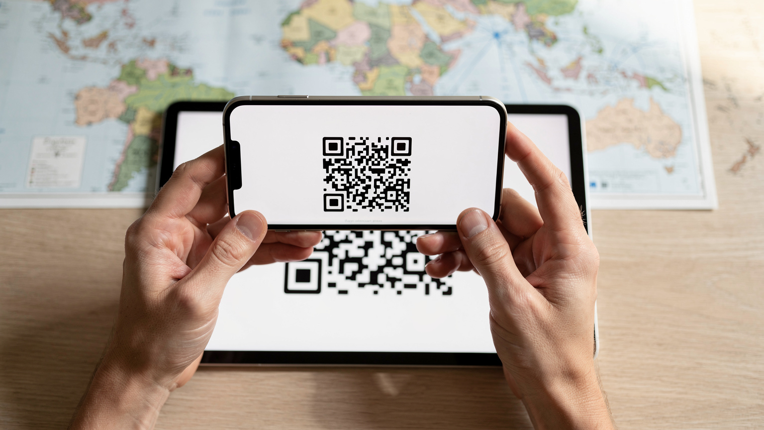 A close-up shot of hands holding a smartphone, scanning a QR code displayed on a tablet, with a blurred travel map in the background.