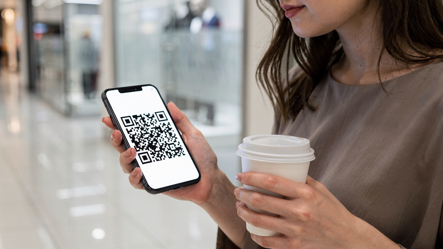 Bir alışveriş merkezinde elinde bir karton kahveyle yürüyen bir kadının cep telefonunu yakın çekim görüntüsü. Ekranında bir eSIM kurulumu için QR kodu açılış sayfası görünüyor, yüzünde hafif bir endişe var.