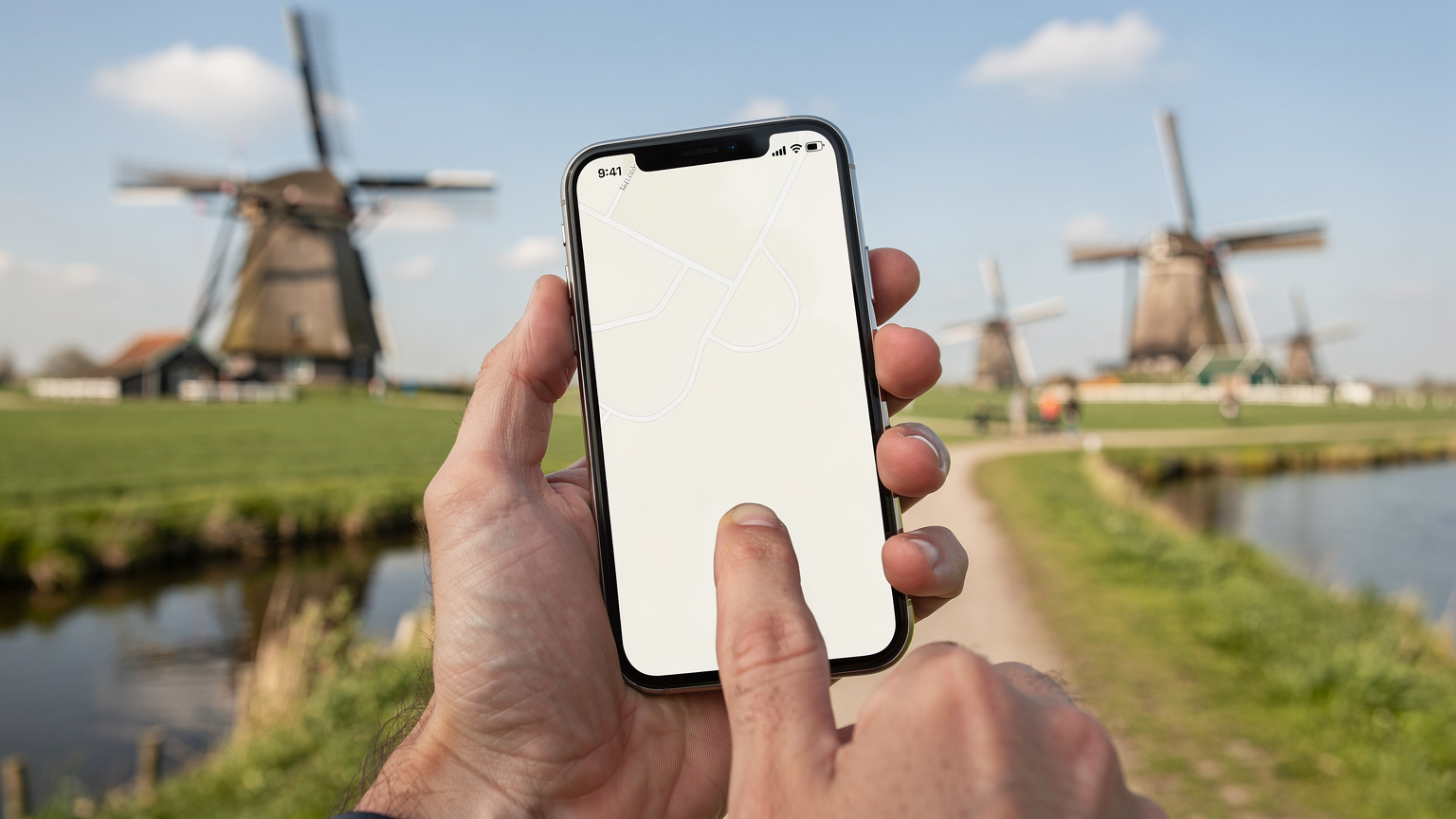 Hollanda'nın kırsalında, geleneksel yel değirmenlerinin ve yemyeşil tarlaların arasında, bir el akıllı telefon ekranında harita uygulamasını kontrol ediyor.