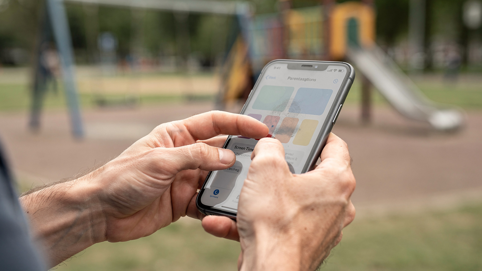 Bir ebeveynin elleri, bir akıllı telefon ekranında 'Ekran Süresi' ayarlarını kontrol ediyor, arka planda bulanık bir çocuk parkı görünüyor.