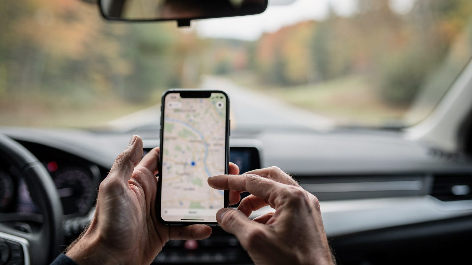 A person's hands are shown clearly holding a smartphone, interacting with a map app to find directions while driving, with a subtle blur of the road in the background.