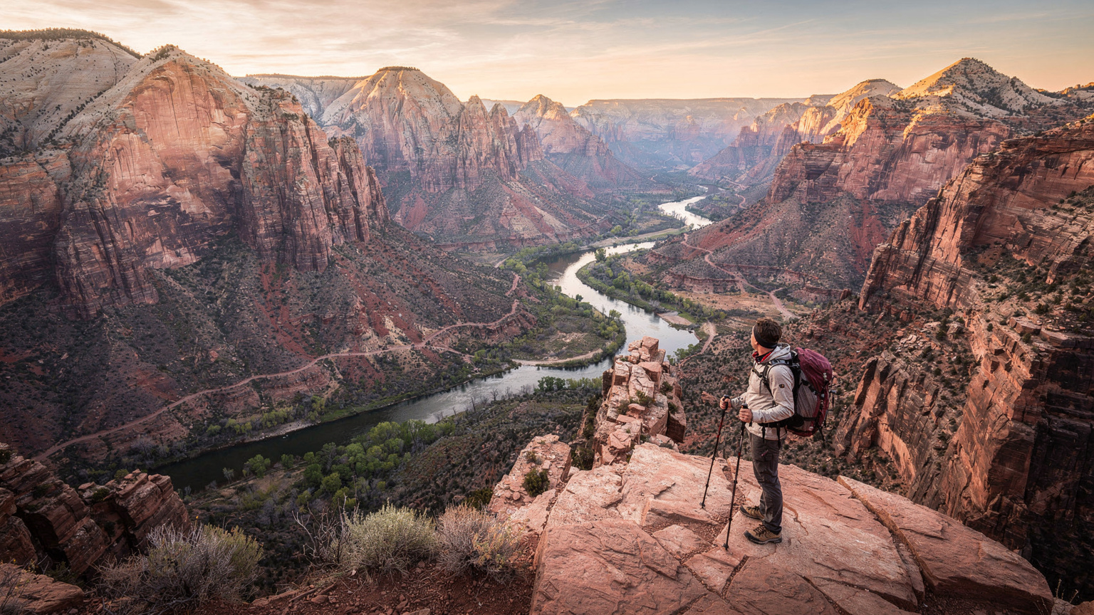 A lone hiker stands at a scenic overlook in Zion National Park, bathed in the soft light of dawn, gazing out at the vast, red rock landscape.
