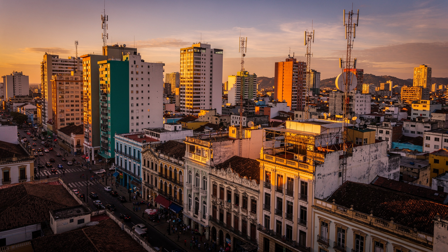 Vista aérea de uma cidade vibrante na América do Sul ao entardecer, com edifícios altos e coloridos misturados com arquitetura histórica, e várias antenas de celular visíveis nos telhados, iluminadas pelo sol poente.