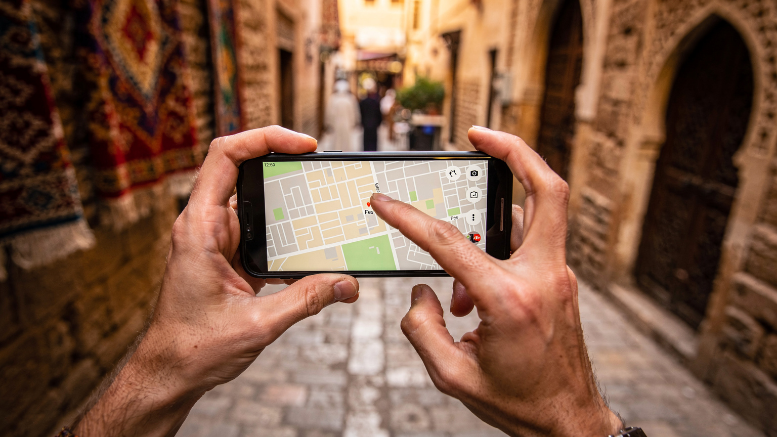 Nahaufnahme eines Smartphones in den Händen eines Reisenden, der eine Karte in der Medina von Fes, Marokko, betrachtet, wobei die Geste des Navigierens im Vordergrund steht.