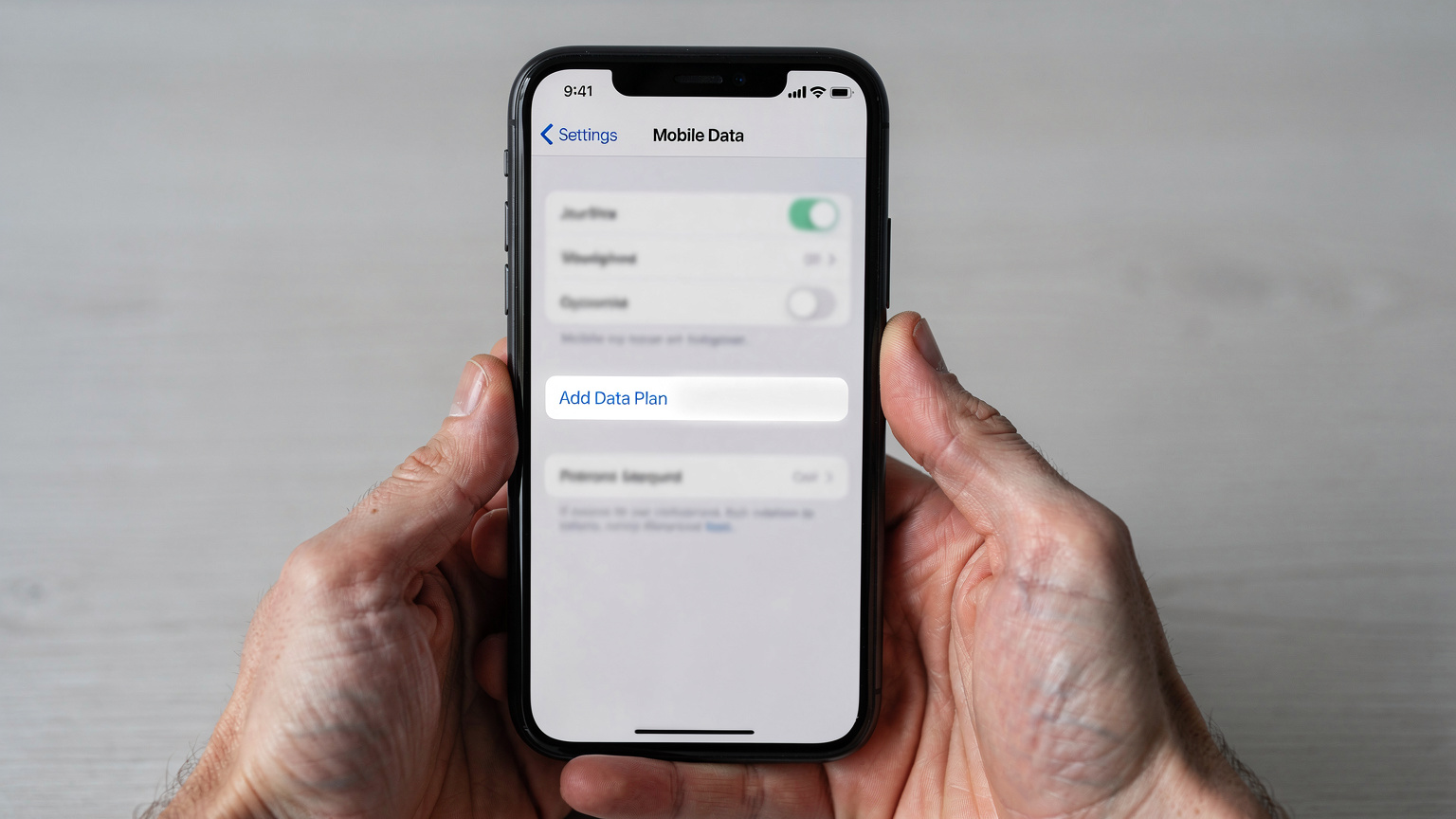 Primer plano de unas manos sosteniendo un smartphone, mostrando la sección de ajustes de 'Datos móviles' con la opción 'Añadir plan de datos móviles' en un iPhone moderno.