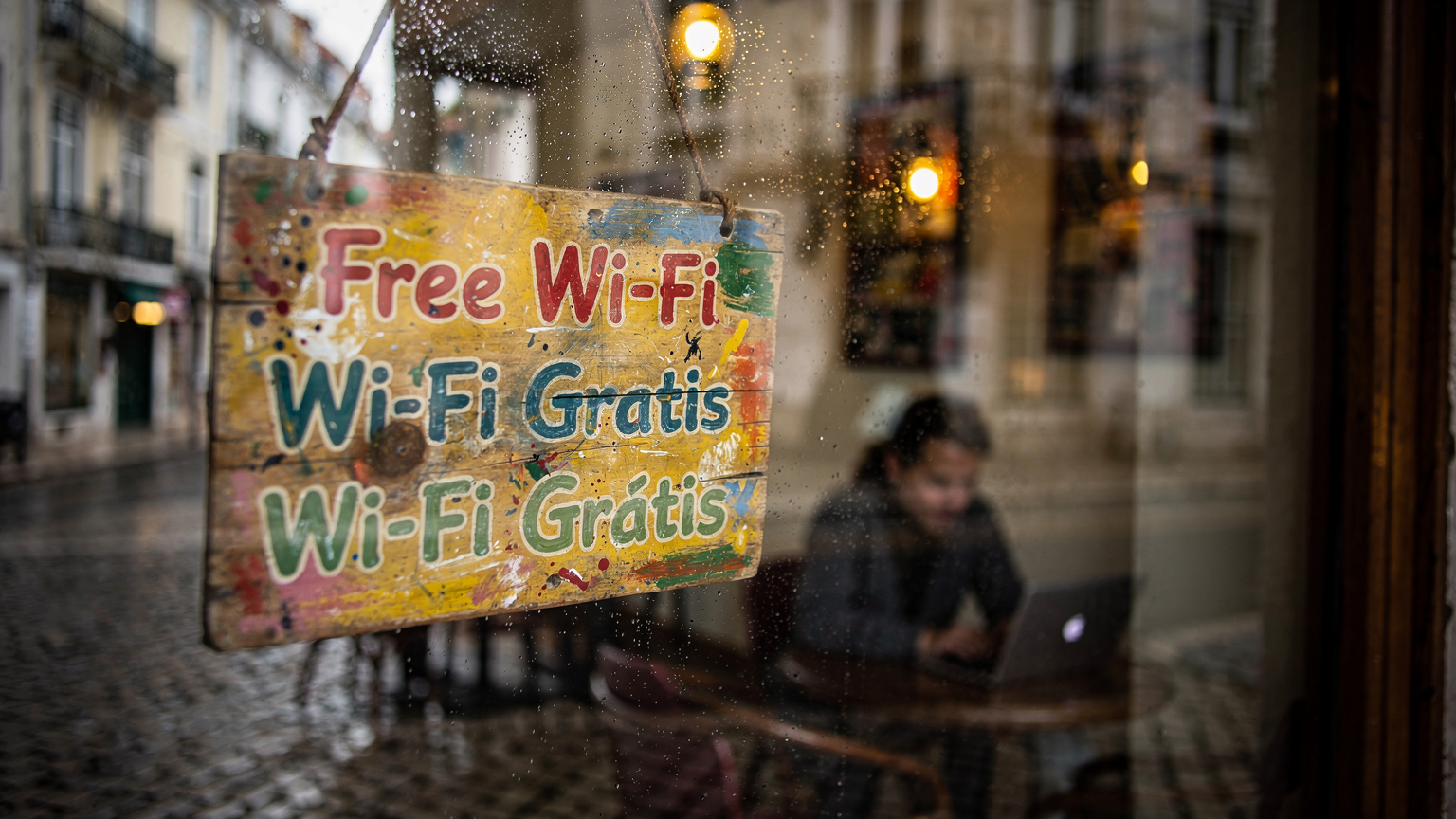 Un cartello di un caffè con la scritta "Wi-Fi gratuito" in diverse lingue, visto attraverso una finestra appannata, con la silhouette di una persona seduta con il suo laptop all'interno.