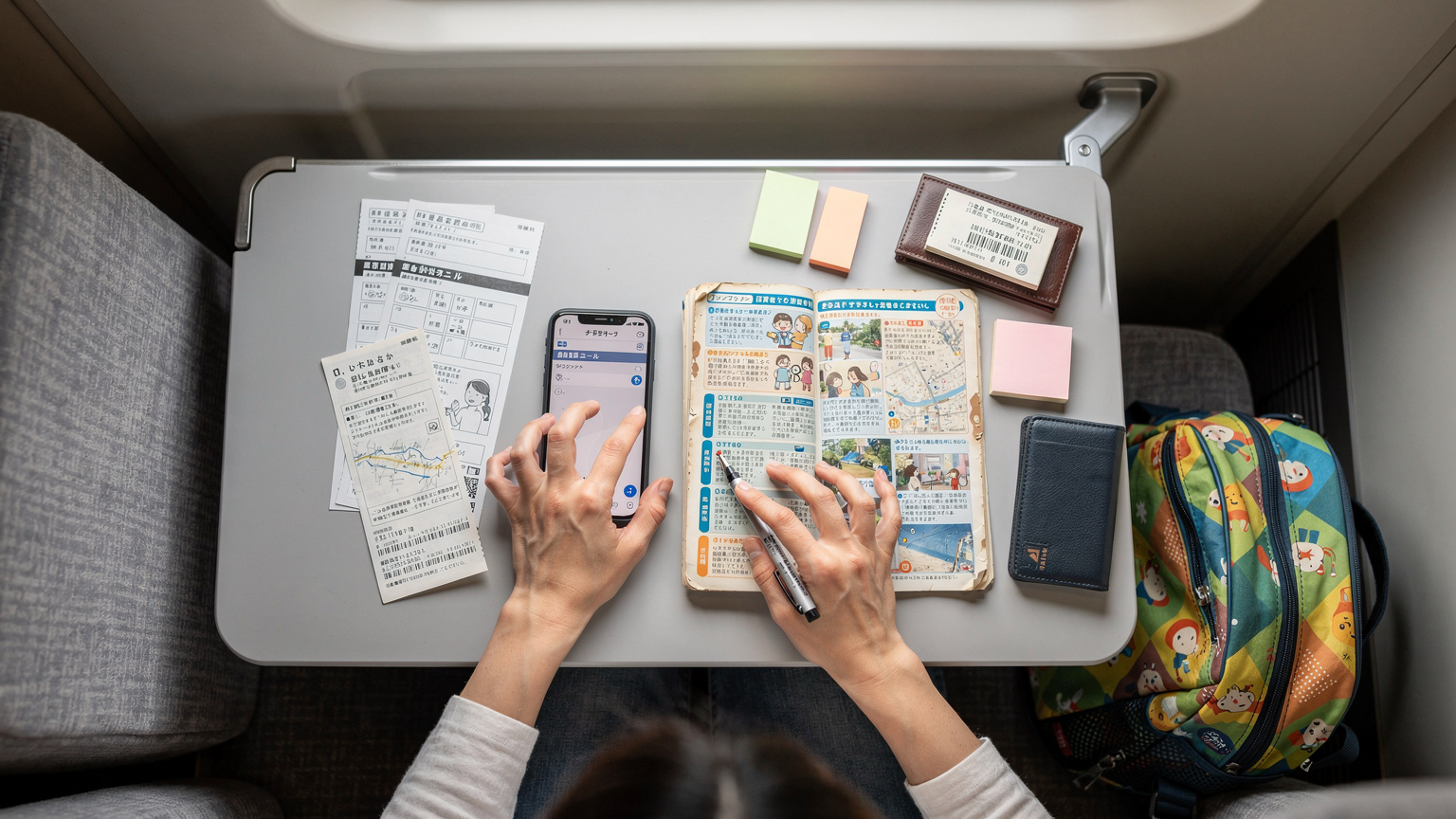 日本の電車内で、旅行ガイドブックとスマートフォンをテーブルに置き、家族旅行の計画を最終確認している母親の俯瞰ショット。隣には子どものリュックサックが見える。