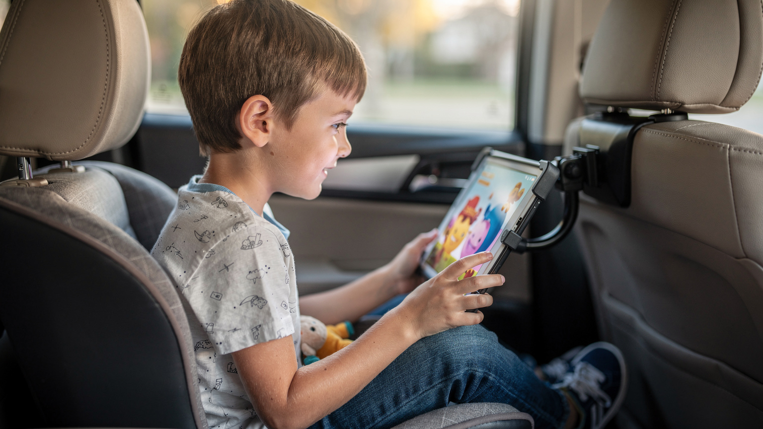 A young boy in the back seat of a car laughing and watching a cartoon on a tablet, safely secured in a car seat.
