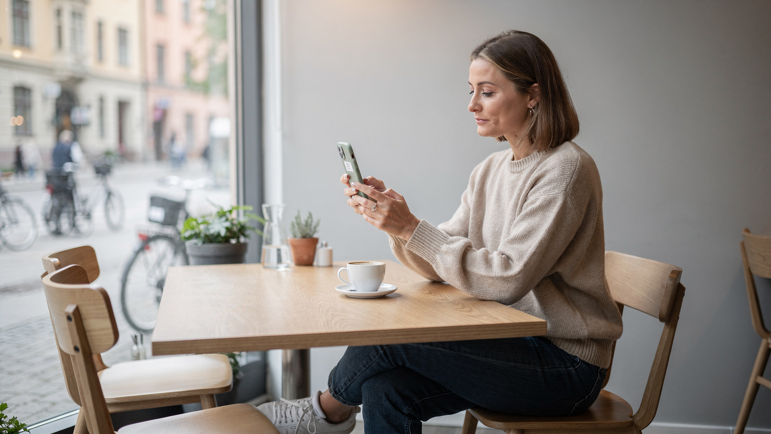 En kvinna sitter vid ett modernt cafébord i Stockholm, håller upp sin nya iPhone och är redo att skanna en QR-kod från sin operatörs hemsida för att aktivera sitt eSIM.