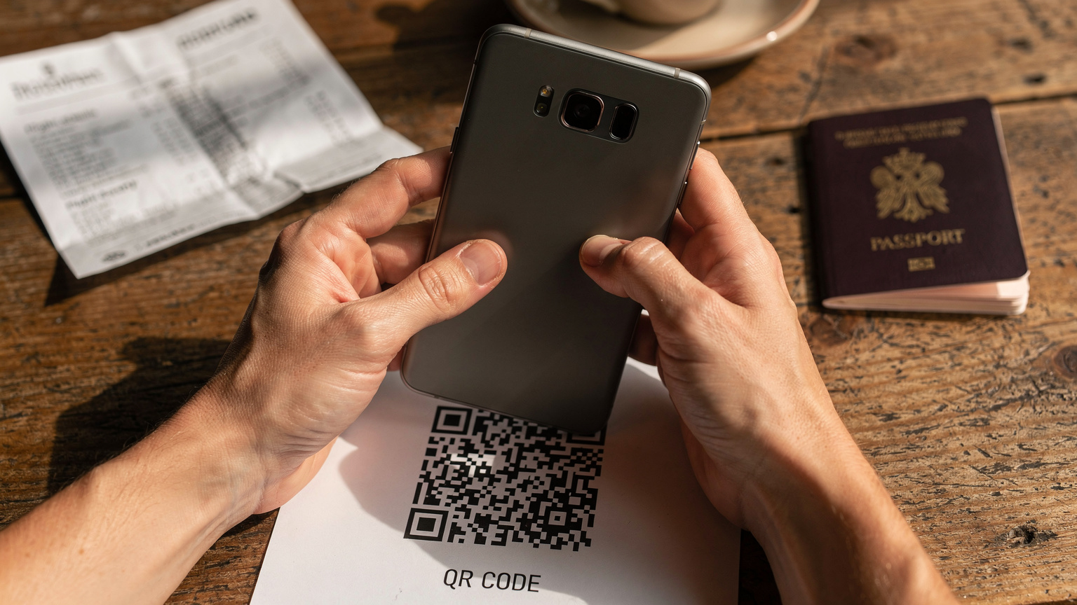 Närbild på ett par händer som håller en Android-telefon och noggrant skannar en QR-kod för eSIM-aktivering, med ett pass och en resplan på ett kafébord i bakgrunden.