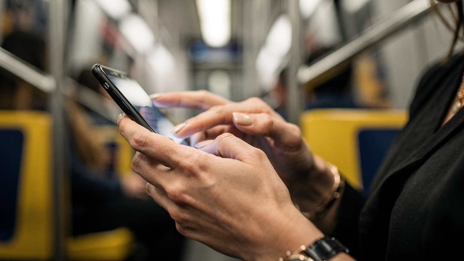 Detailní záběr rukou cestovatelky, která drží chytrý telefon a prochází nastavením mobilních dat, s rozostřeným pozadím interiéru newyorského metra.