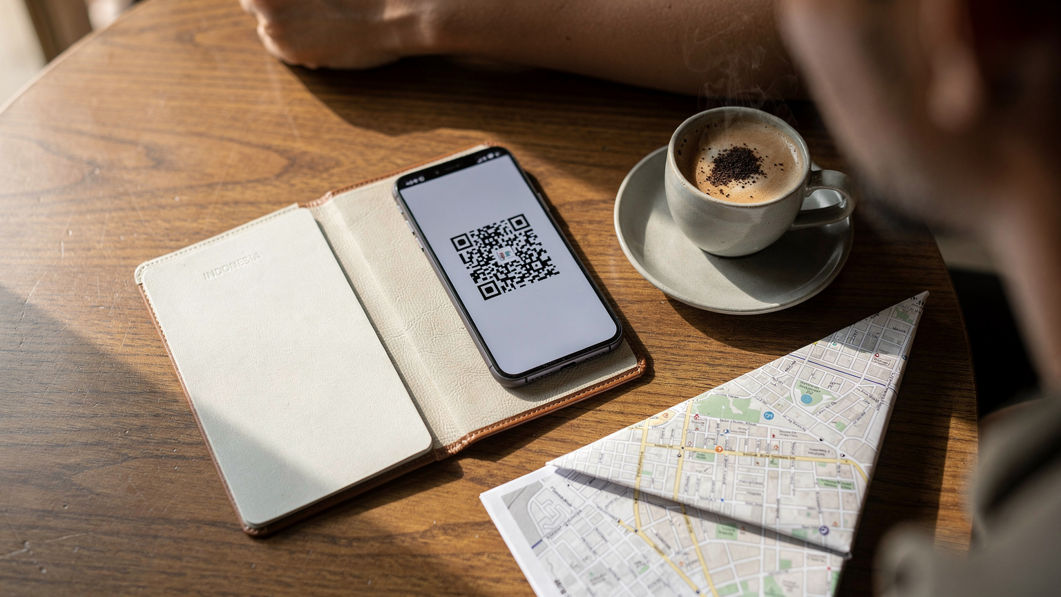 Tampilan overhead dari meja kafe yang rapi dengan paspor terbuka, ponsel yang menampilkan kode QR eSIM, kopi panas, dan selembar peta kota yang sebagian terlihat.