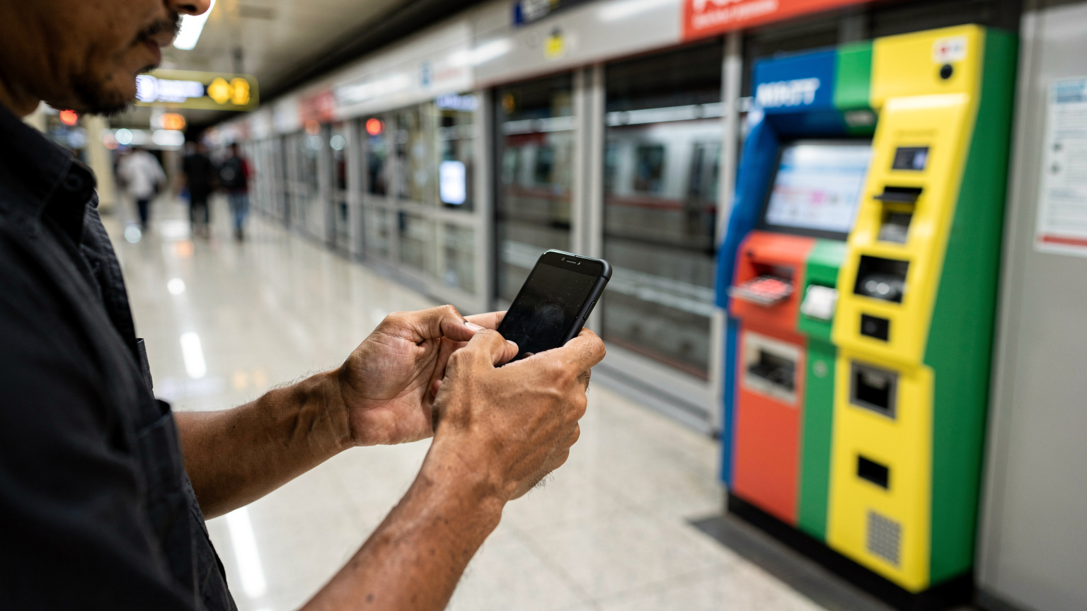 Tangan seorang pria sedang memegang ponsel di dekat mesin tiket otomatis berwarna-warni di stasiun MRT Jakarta, menunjukkan momen interaksi dengan teknologi dalam perjalanan.