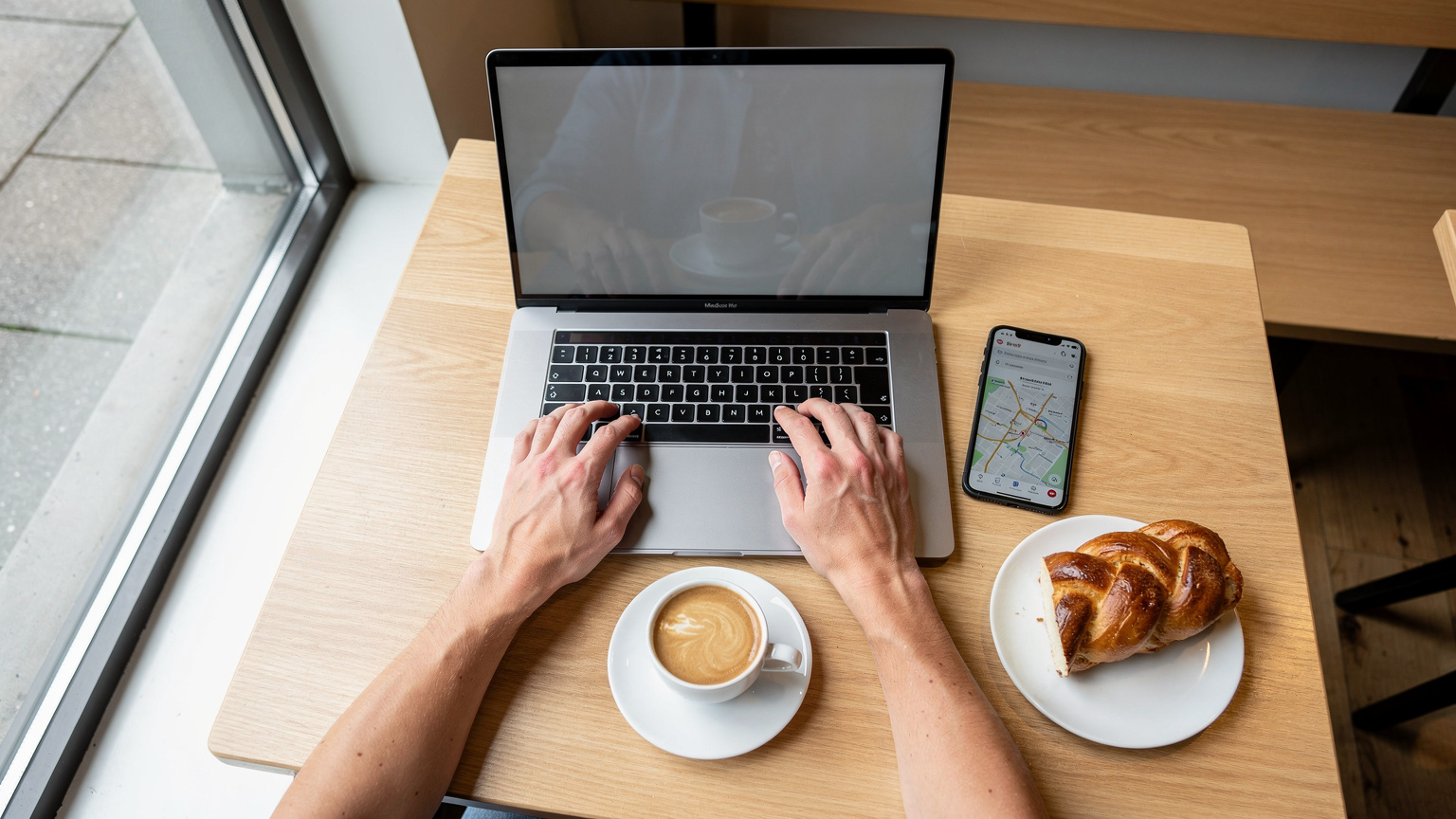 O persoană lucrează la laptop într-o cafenea modernă din Zurich, cu o ceașcă de cafea și o prăjitură elvețiană pe masă, telefonul afișând o hartă.