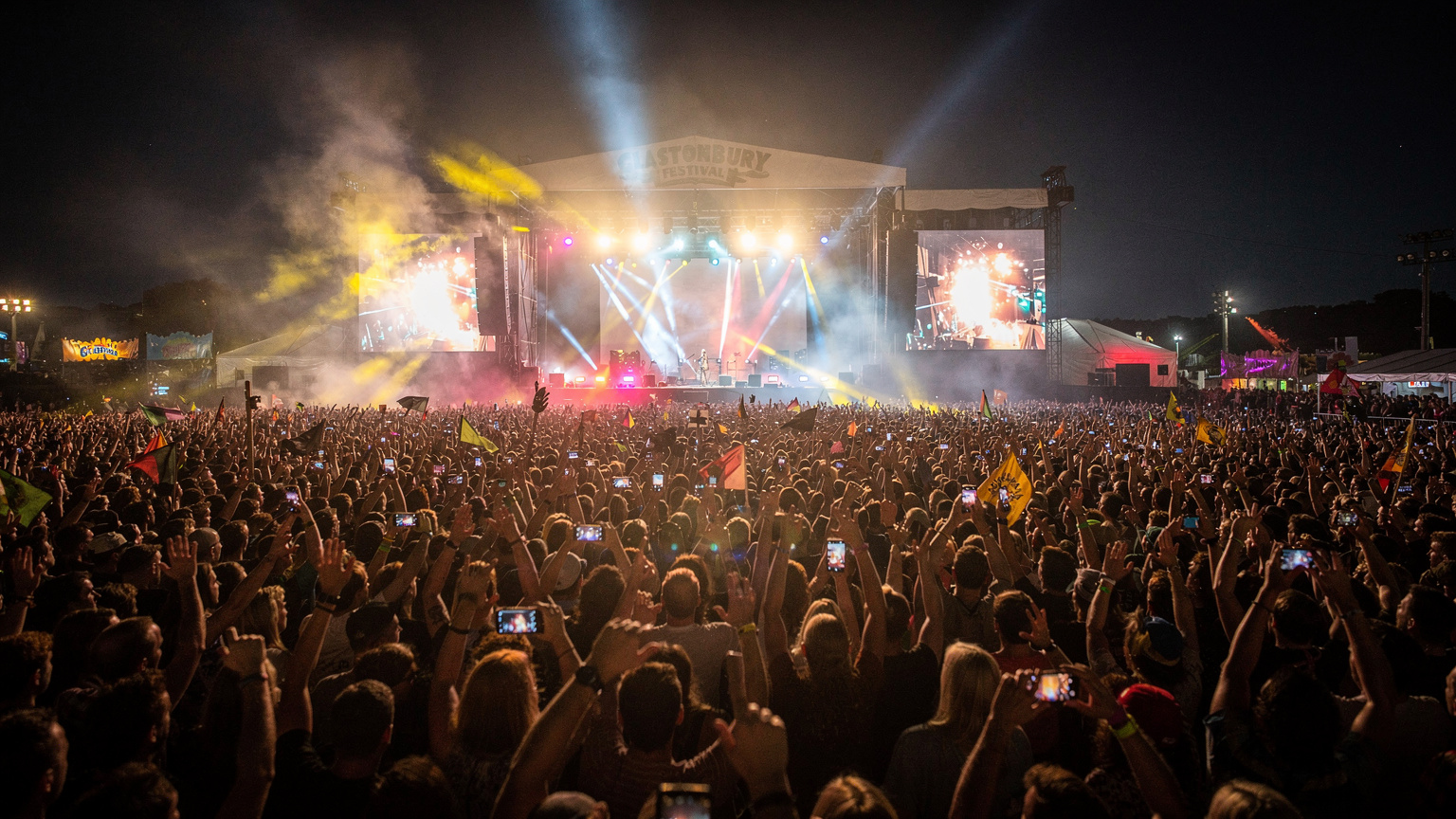 Glastonbury Festivali'nde devasa bir sahnenin önünde toplanmış, havaya ellerini kaldırmış binlerce insan, ışıklandırmalar ve sis efektleri eşliğinde müziğin keyfini çıkarıyor. Gecenin karanlığı ve sahnenin parlak ışıkları altında kalabalığın enerjisi hissediliyor.