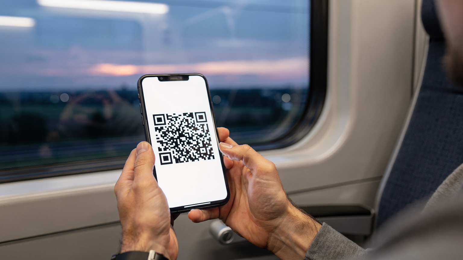 Close-up of a traveler's hands holding a smartphone, displaying a QR code for eSIM activation against a softly blurred background of a European train window at dusk.