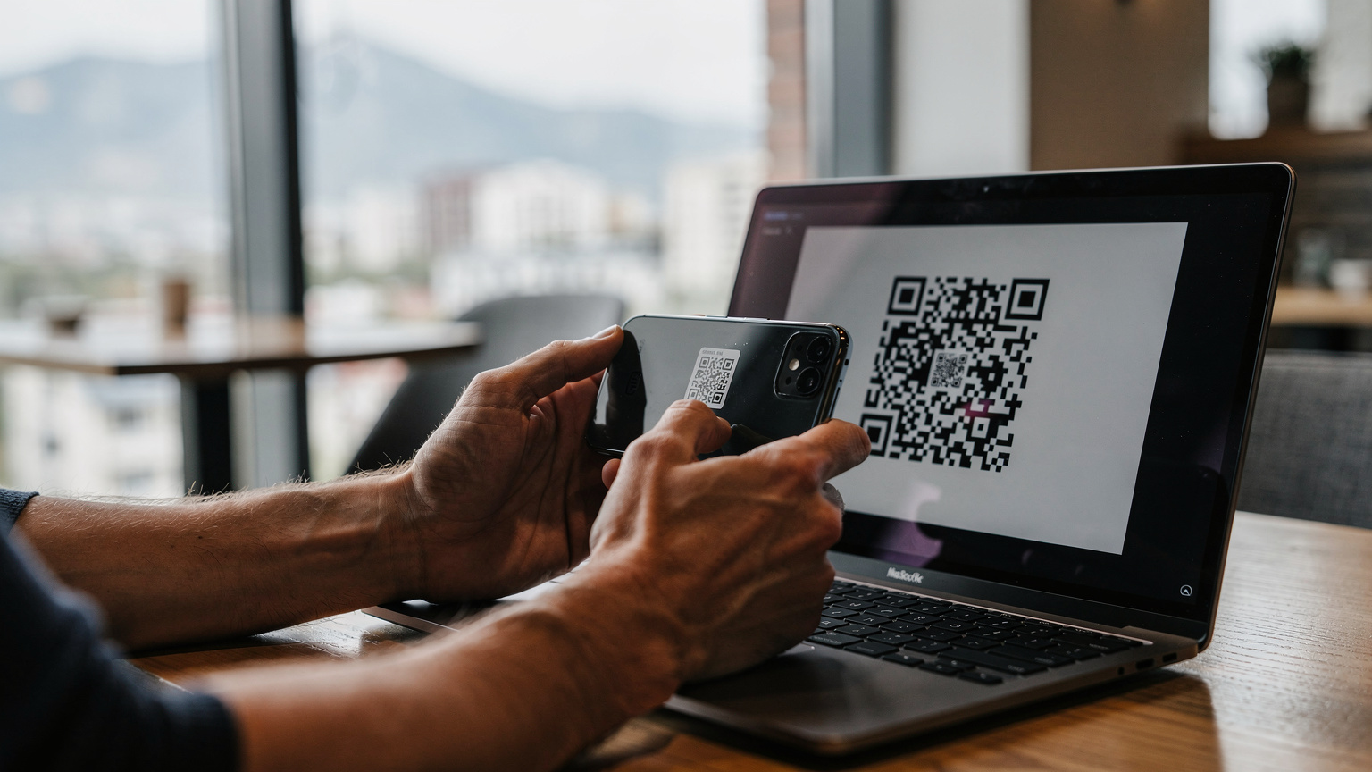 A digital nomad's hands holding a smartphone, scanning an eSIM QR code displayed on a laptop screen, in a well-lit, minimalist co-working cafe setting in Almaty.