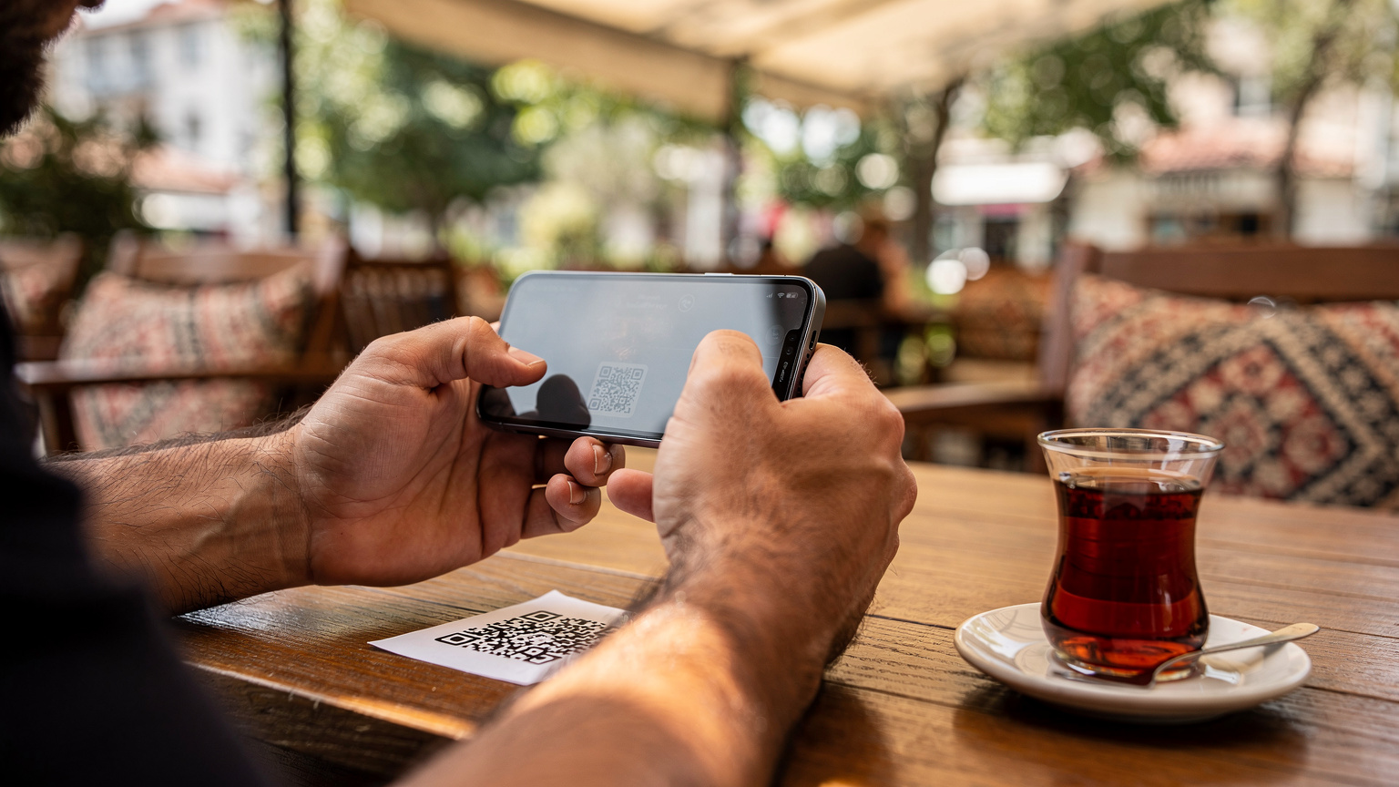 Мъж сканира QR код от разпечатан документ с камерата на своя iPhone 18, докато седи на маса в традиционно турско кафене в квартал Доганджилар, Истанбул, с чаша чай до него.