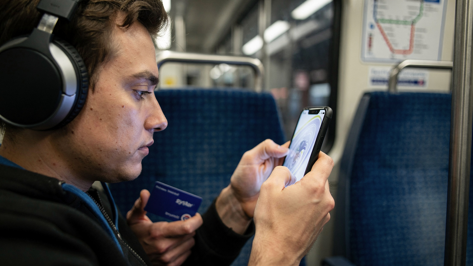 Пътешественик в метрото на Лондон държи своя iPhone 18 и разглежда менюто за настройки на eSIM, докато слуша музика със слушалки и държи карта Oyster.