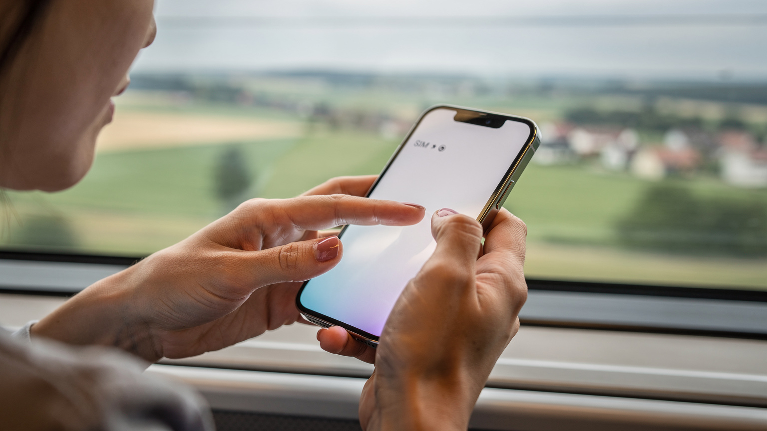 ヨーロッパの高速鉄道（ドイツのICE）の窓際に座り、走行中の車窓を背景にiPhoneのeSIM設定画面を操作している日本人女性の手元。
