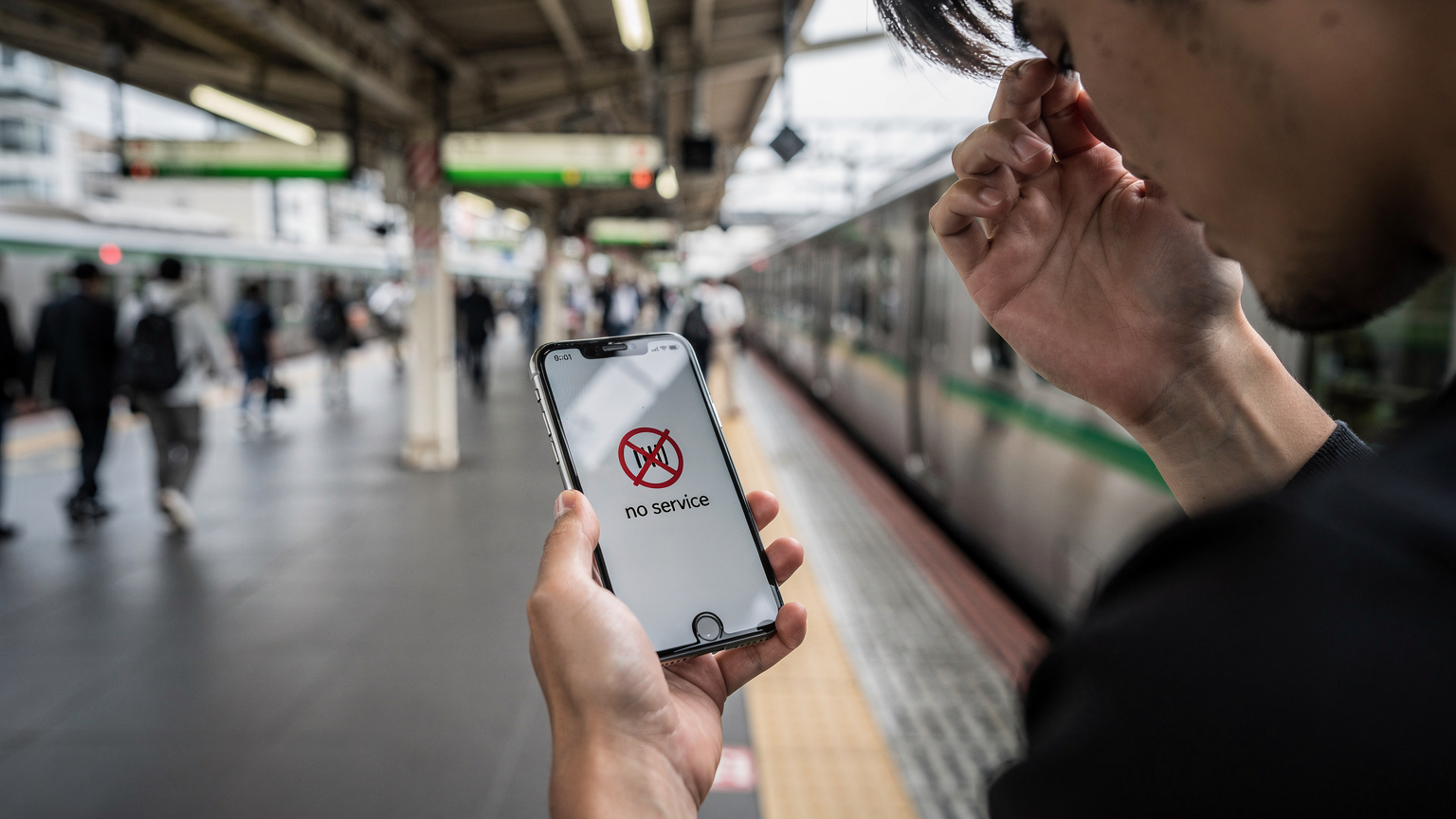 A traveler's hands are shown, one holding an iPhone displaying a 'No Service' error, the other rubbing their temple in frustration, set against the blurry background of a bustling train station platform in Tokyo's Yamanote line.