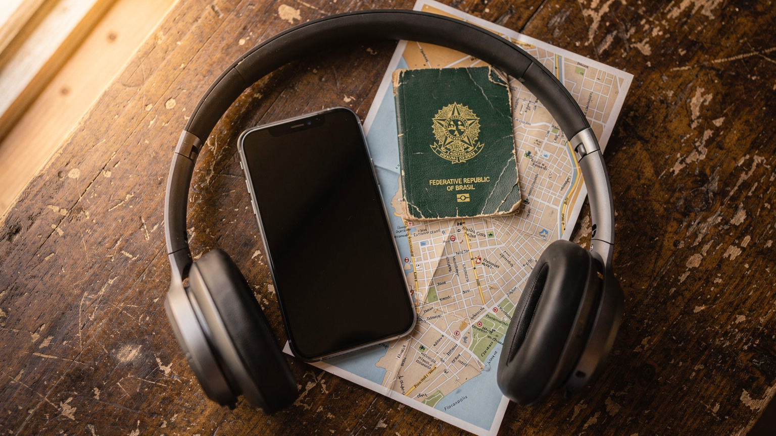 An editorial flat-lay shot on a rustic wooden table featuring a modern iPhone, a passport from a South American country, noise-canceling headphones, and a small, folded paper map, evoking a sense of prepared travel.