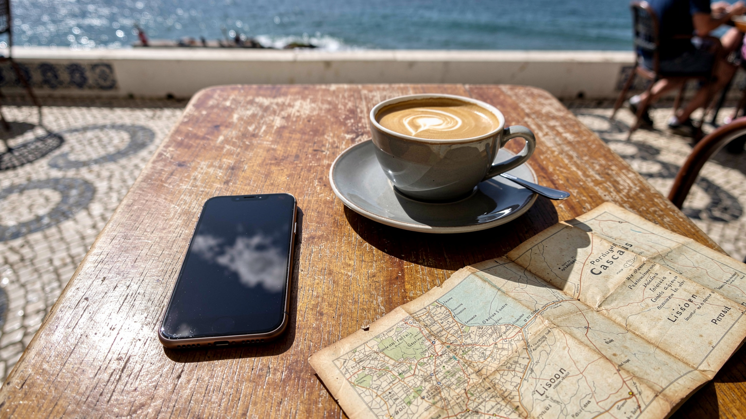 Uma vista aérea de uma mesa de café numa esplanada em Cascais, Portugal, com um iPhone 18, um copo de café, e um mapa turístico de Portugal parcialmente aberto, com o Oceano Atlântico ao fundo.