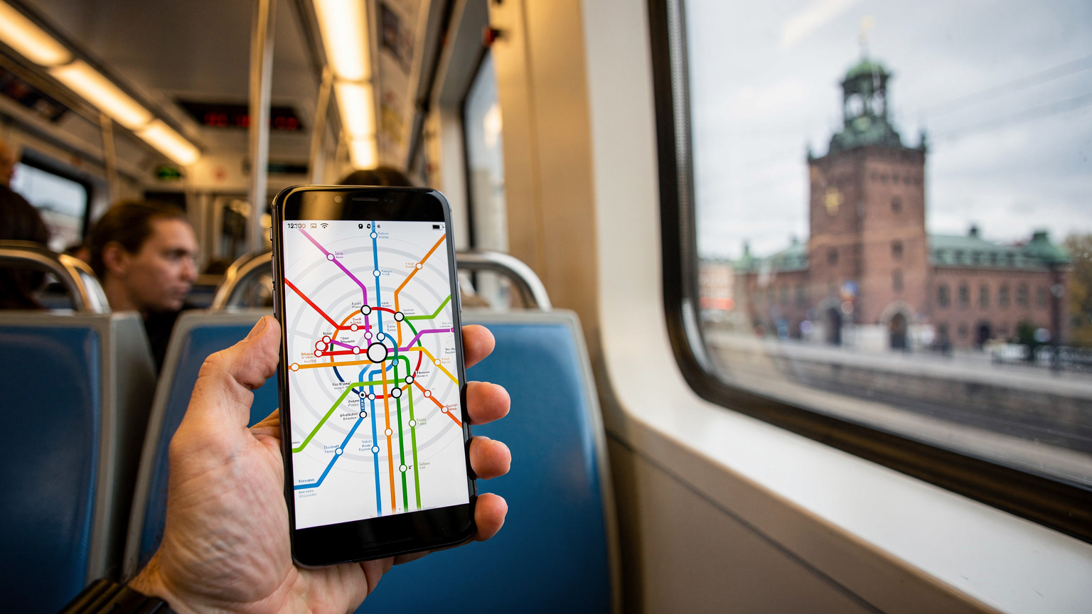En karta över Stockholms tunnelbana visas på en smartphoneskärm, hållen av en hand i en tunnelbanevagn, med utsikt över Stadshuset genom fönstret.
