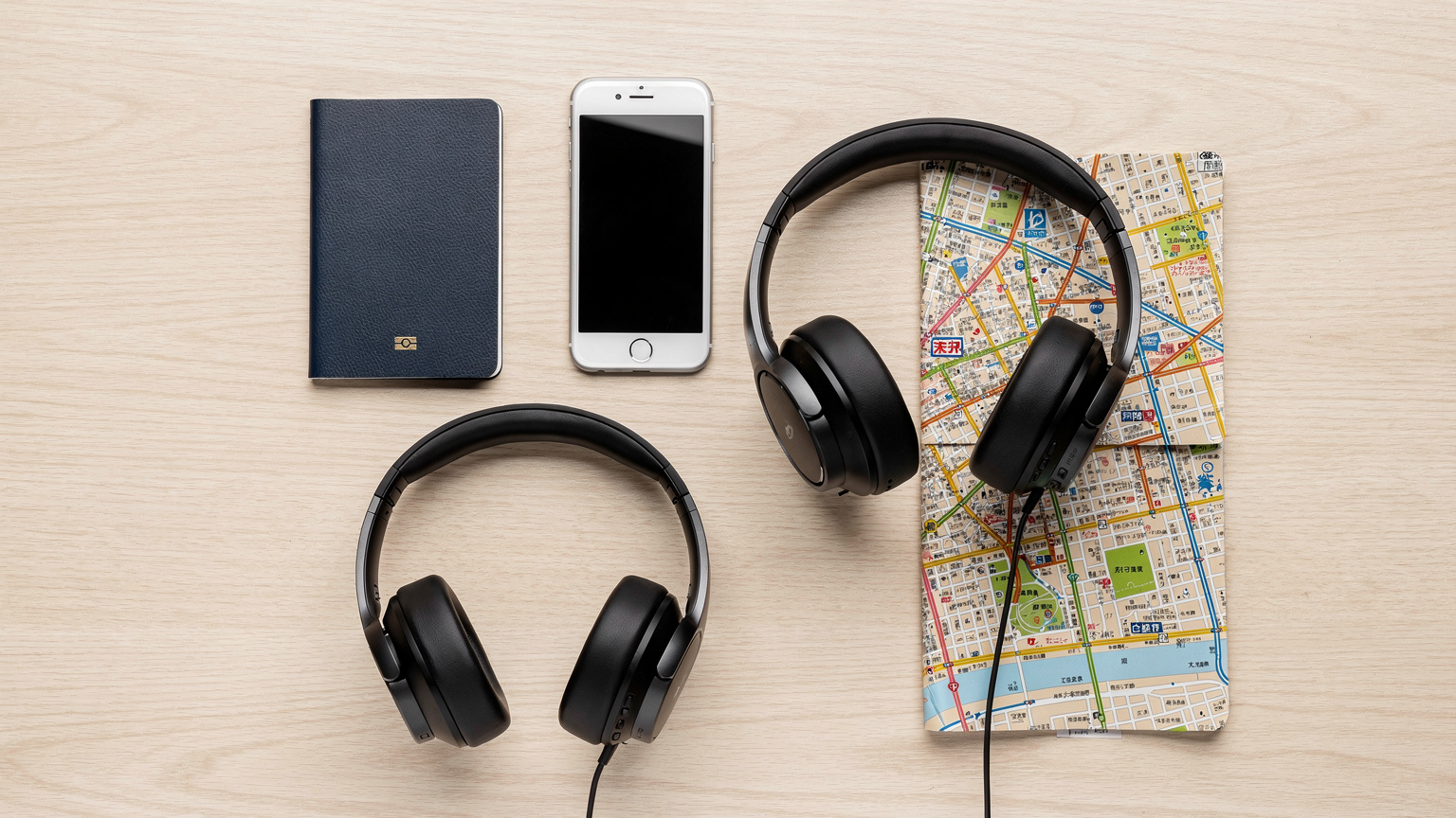 An overhead flat-lay of travel essentials: a passport, a smartphone, noise-cancelling headphones, and a neatly folded map of Tokyo, arranged on a light wooden table.