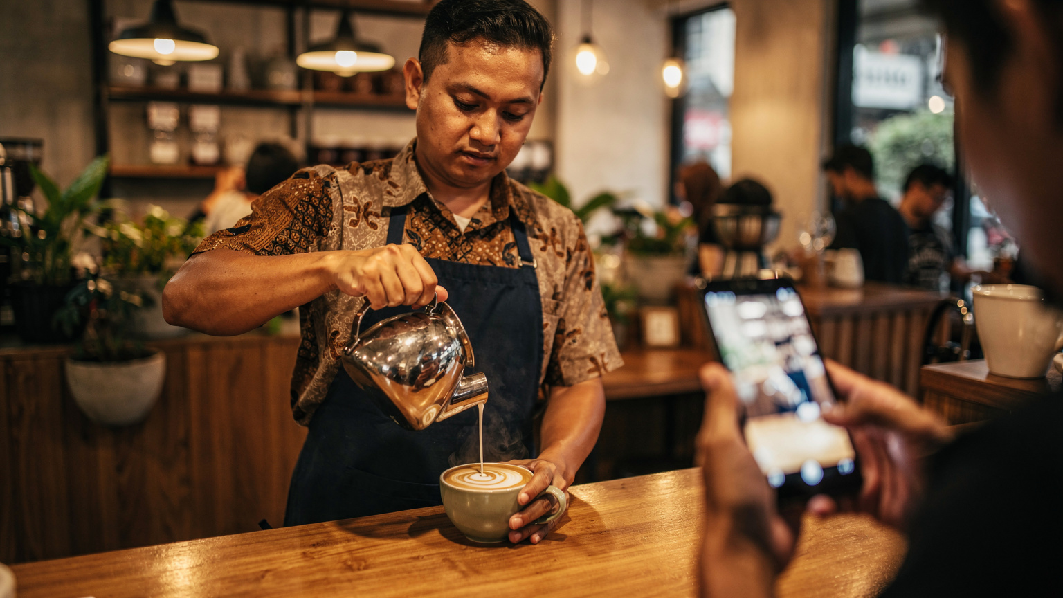 Seorang barista di kafe modern di Jakarta menyajikan kopi, dengan ponsel yang menampilkan pengaturan WiFi di latar belakang, mencerminkan kebutuhan koneksi di lingkungan perkotaan yang sibuk.