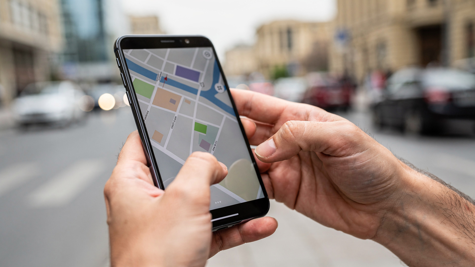 A close-up shot of a traveler's hands, specifically their thumbs, swiftly navigating a map application on a smartphone. The phone is a modern model, with no visible logos or UI text, emphasizing the tactile interaction and focus on navigation. The background is a slightly blurred, vibrant street scene in Baku, suggesting movement and exploration.