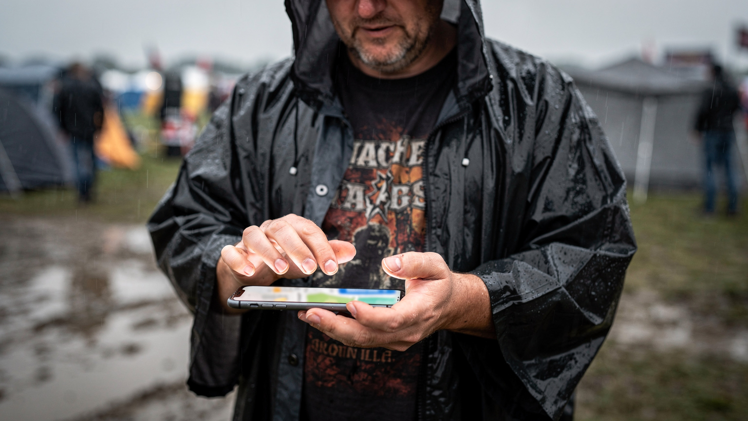 Ein Festivalbesucher im Regenmantel navigiert auf seinem Smartphone durch eine Karte, während im Hintergrund ein regnerisches Festivalgelände mit Zelten und Menschen zu sehen ist.