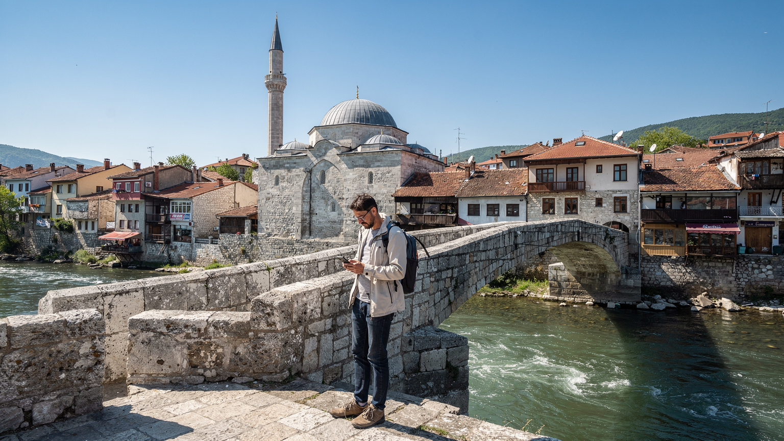 Prizren'in eski köprüsünde, tarihi camii ve Bistrica Nehri'nin arka planında, akıllı telefonundan harita kontrol eden bir gezgin.