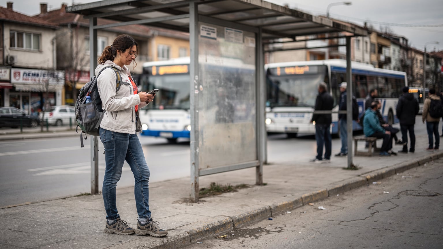 Priştine'deki otobüs durağında, otobüs saatlerini kontrol etmek için telefonuna bakan genç bir gezgin, arka planda yerel otobüsler görünüyor.
