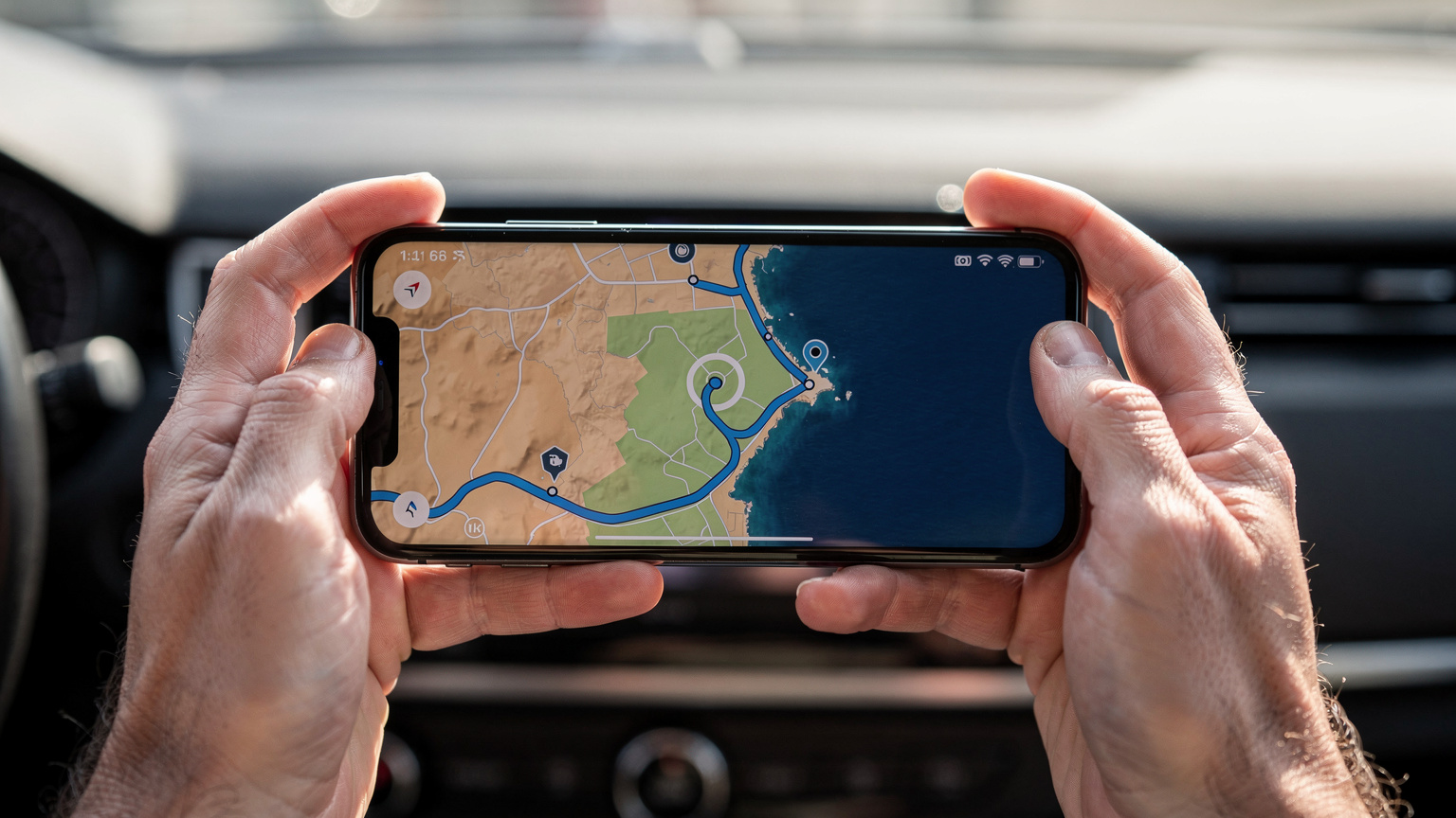 Een close-up van twee handen die een smartphone vasthouden. Op het scherm van de telefoon is een Google Maps-route te zien die langs de kustlijn van Spanje loopt, met markeringen voor stops en de huidige locatie van de gebruiker.