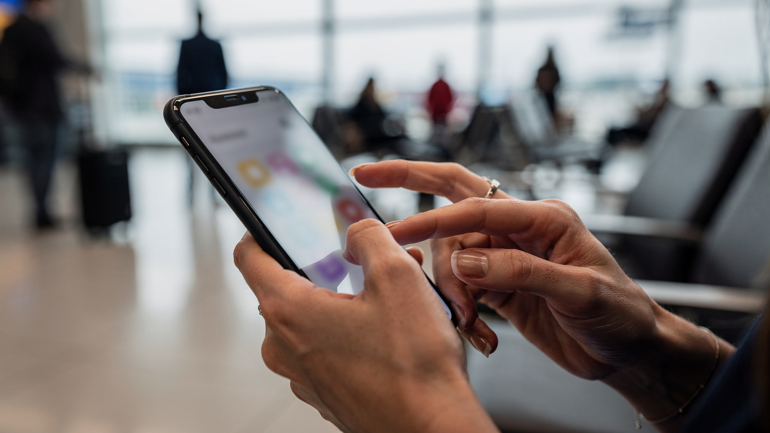 Nahaufnahme der Hände einer Mutter, die auf einem iPhone die Bildschirmzeit-Einstellungen für das Gerät ihres Kindes anpasst, mit einem belebten Flughafen im unscharfen Hintergrund.