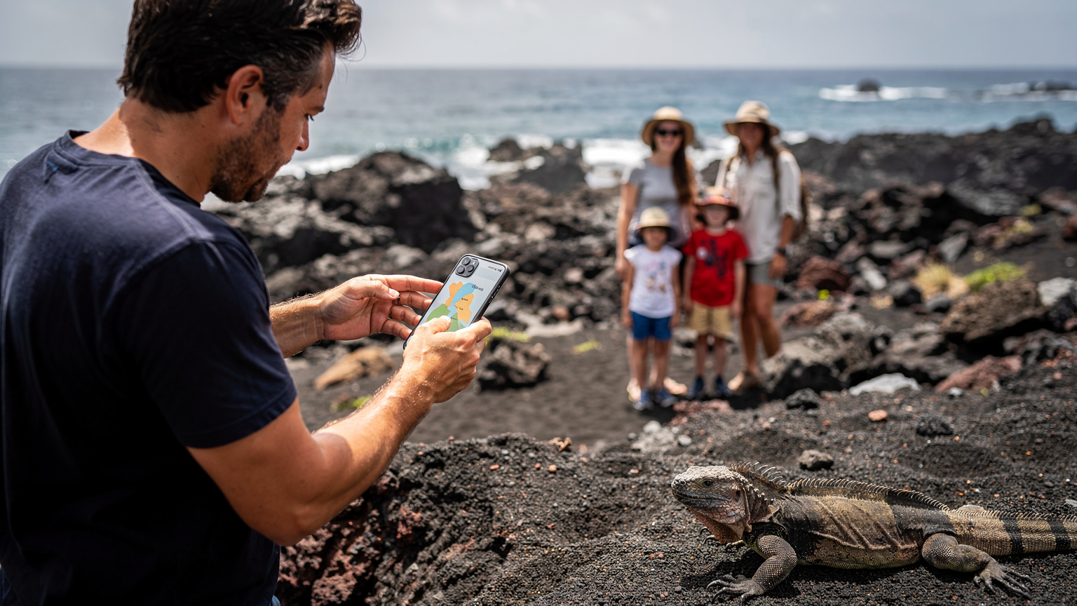 Un padre, con su familia de fondo, consulta un mapa offline en su teléfono mientras observan una iguana marina en una playa de arena volcánica en las Galápagos. El paisaje es rocoso y árido, con el océano Pacífico al fondo.