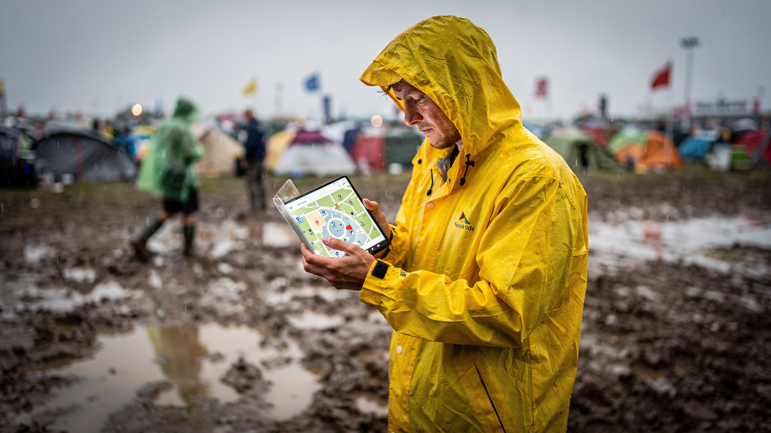 En festivalgænger i regntøj og gummistøvler studerer et offline kort over Roskilde Festival på sin smartphone, mens regnen drypper, og mudderet er tydeligt på jorden.