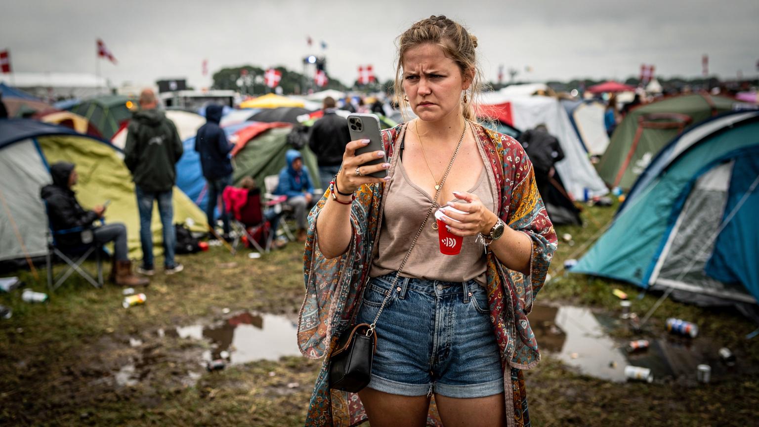 En ung festivalgænger holder sin telefon op for at fange et billede af scenen ved solnedgang på Roskilde Festival, med mange andre mennesker i baggrunden.