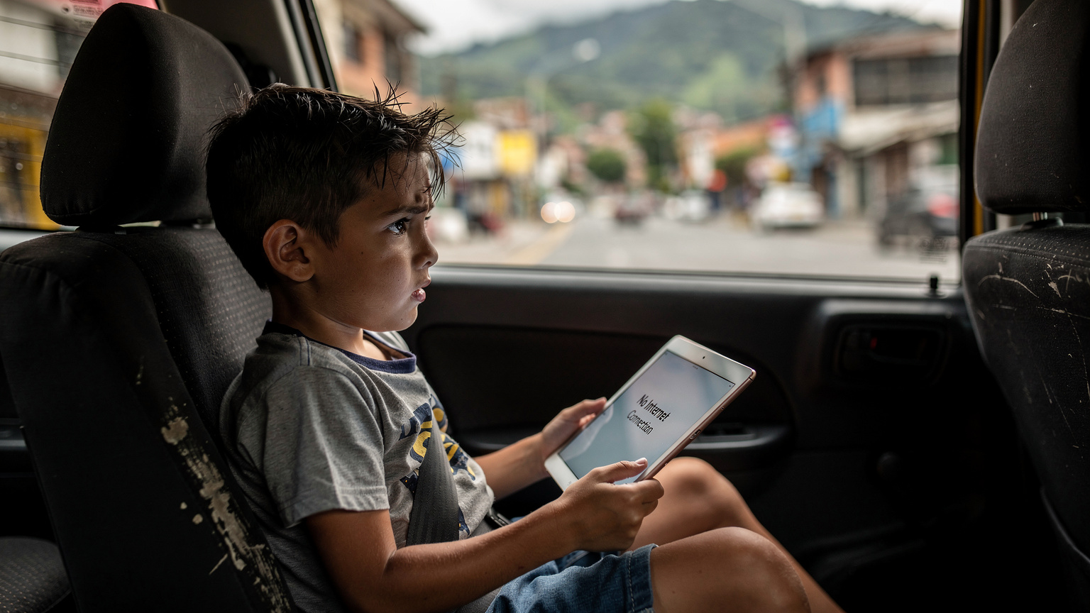 Un niño pequeño, de unos 6 años, está sentado en el asiento trasero de un taxi en Medellín, mirando por la ventana con una expresión de preocupación, mientras su iPad en sus manos muestra una pantalla de 'No hay conexión a internet'.