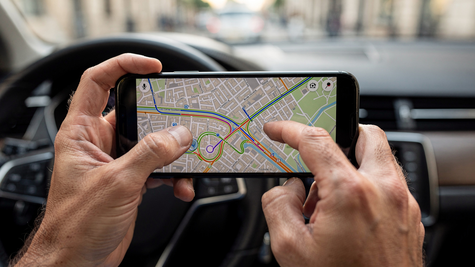 Un primo piano delle mani di una persona che tiene uno smartphone, mostrando un'applicazione di navigazione GPS con un percorso tracciato attraverso una città europea, con l'auto che viaggia su una strada.