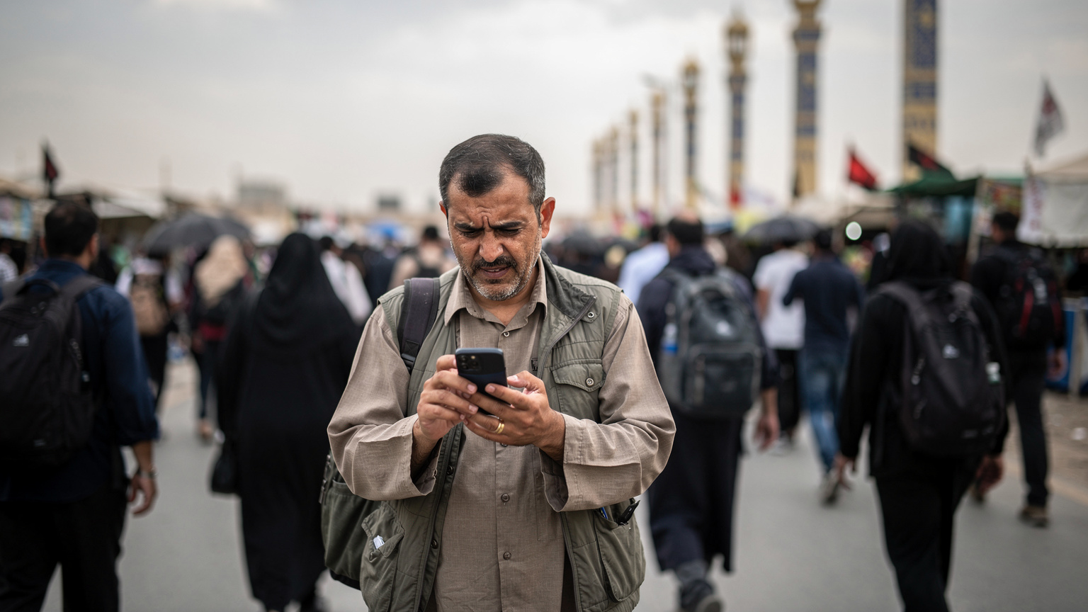 یک زائر در حال تلاش برای پیدا کردن سیگنال تلفن همراه در شلوغی مسیر پیادهروی اربعین، با پسزمینهای از ستونهای نمادین مسیر.