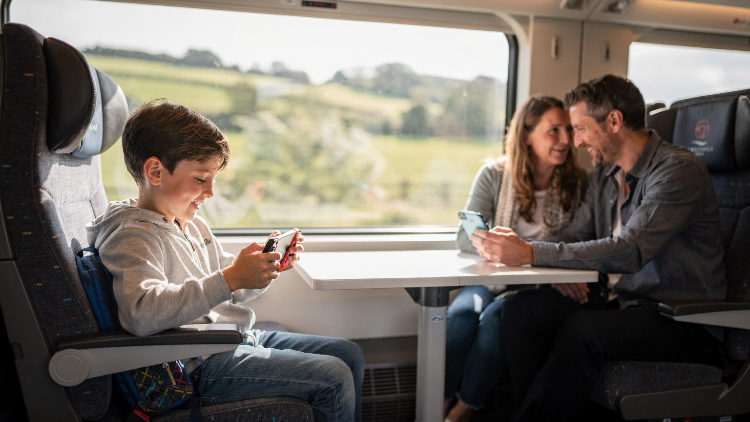 Un niño de unos 9 años, sentado en un asiento de tren moderno en el Reino Unido, absorto en su Nintendo Switch. Sus padres, al fondo, conversan animadamente mientras uno de ellos consulta un mapa en su teléfono, que está conectado a la eSIM.