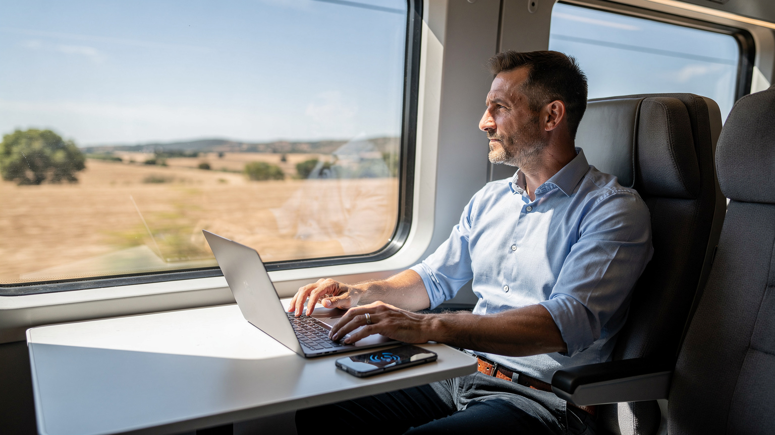 İspanya'da hızlı tren (Renfe) kompartımanında, pencereden dışarıdaki manzarayı izlerken dizüstü bilgisayarında çalışan 40'lı yaşlarında bir erkek.