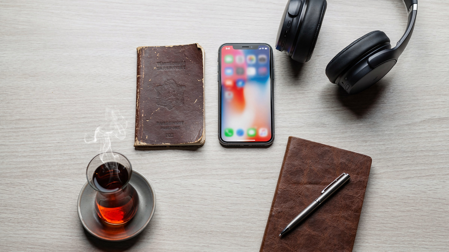 Bir seyahat yazarının çalışma masası üzerinde düzenlenmiş eşyalar: pasaport, açık akıllı telefon (Cellesim uygulaması ekranı), kulaklık, not defteri ve sıcak çay bardağı.