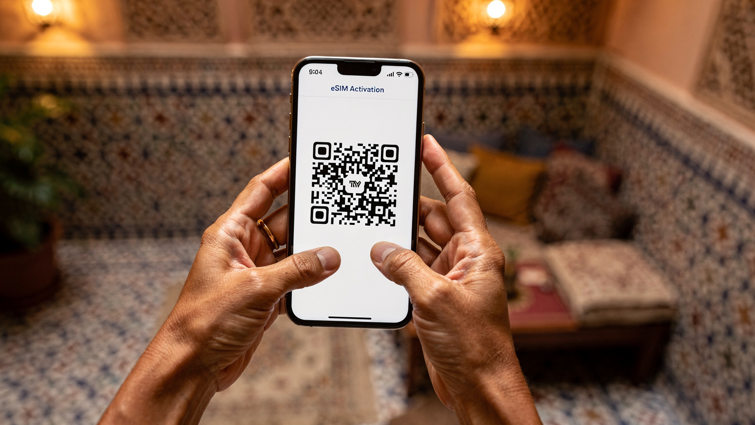 Zbliżenie na dłoń matki trzymającej smartfon, na ekranie którego widać proces aktywacji eSIM z kodem QR. W tle widać rozmyte wnętrze przytulnego, tradycyjnego marokańskiego riadu.