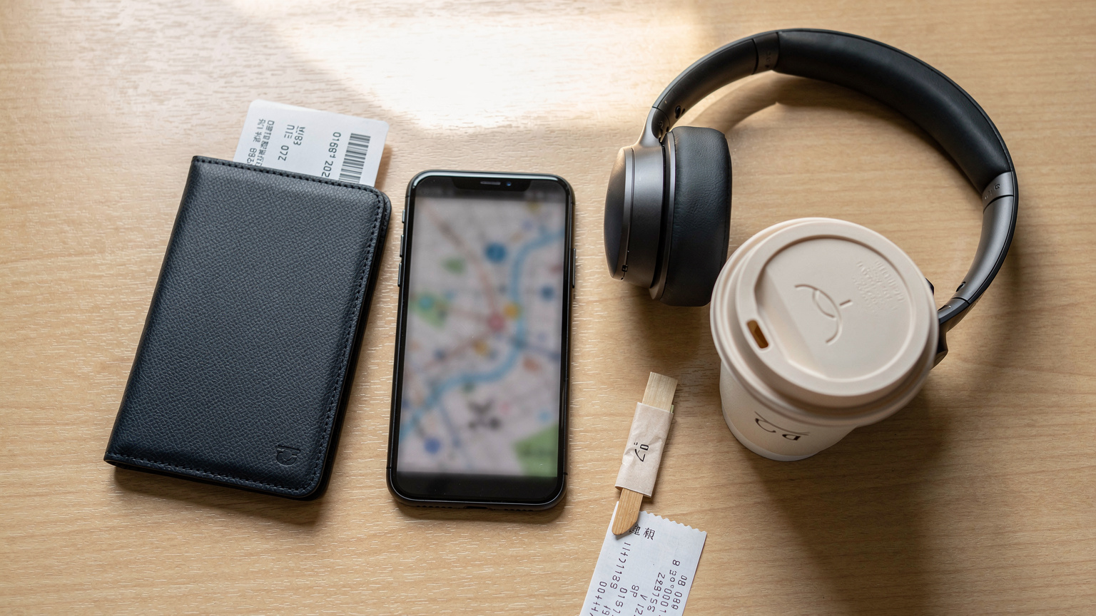 A close-up, overhead flat-lay shot of a traveler's essentials: a passport with a boarding pass tucked inside, a smartphone displaying a blurred map application, noise-canceling headphones, and a small, locally-branded coffee cup from a cafe in Tokyo, Japan.